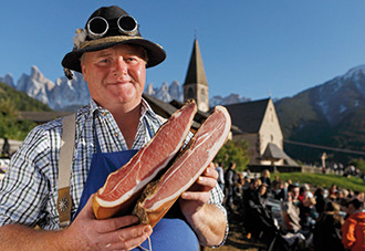 Ein stämmiger Mann hält zwei Südtiroler Schinken in die Kamera